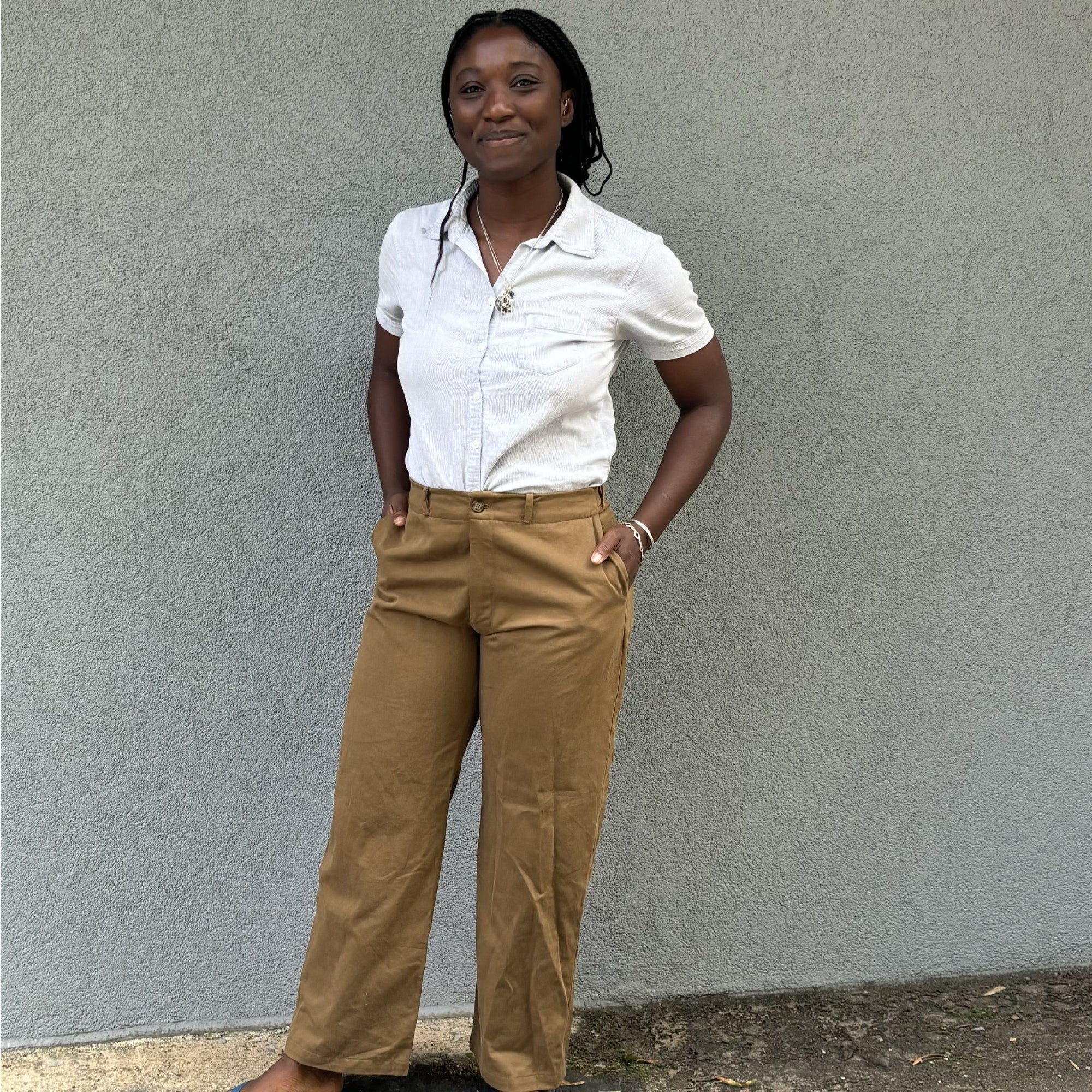 African woman standing and smiling with hand in her olive khaki sanded twill pants in front on a grey wall.