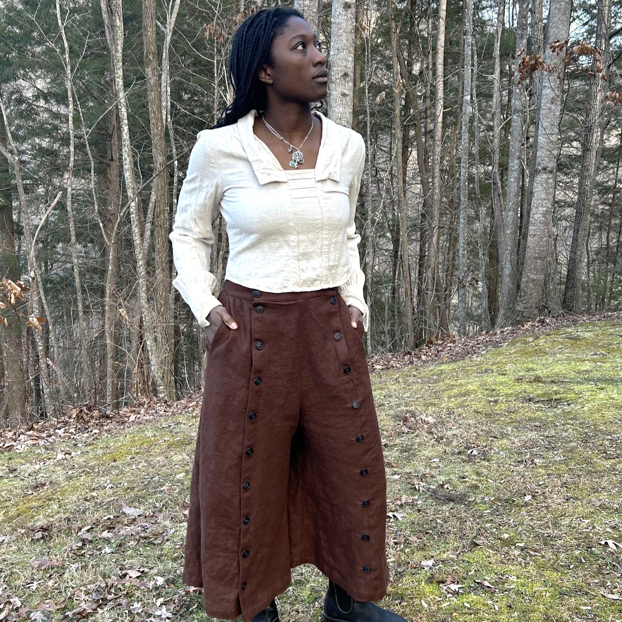 African woman wearing a dark brown linen 231 riding skirt with black buttons in pant form, and a beige 210 armistice blouse. She is looking up and standing outside in the woods with her pockets in her hands.