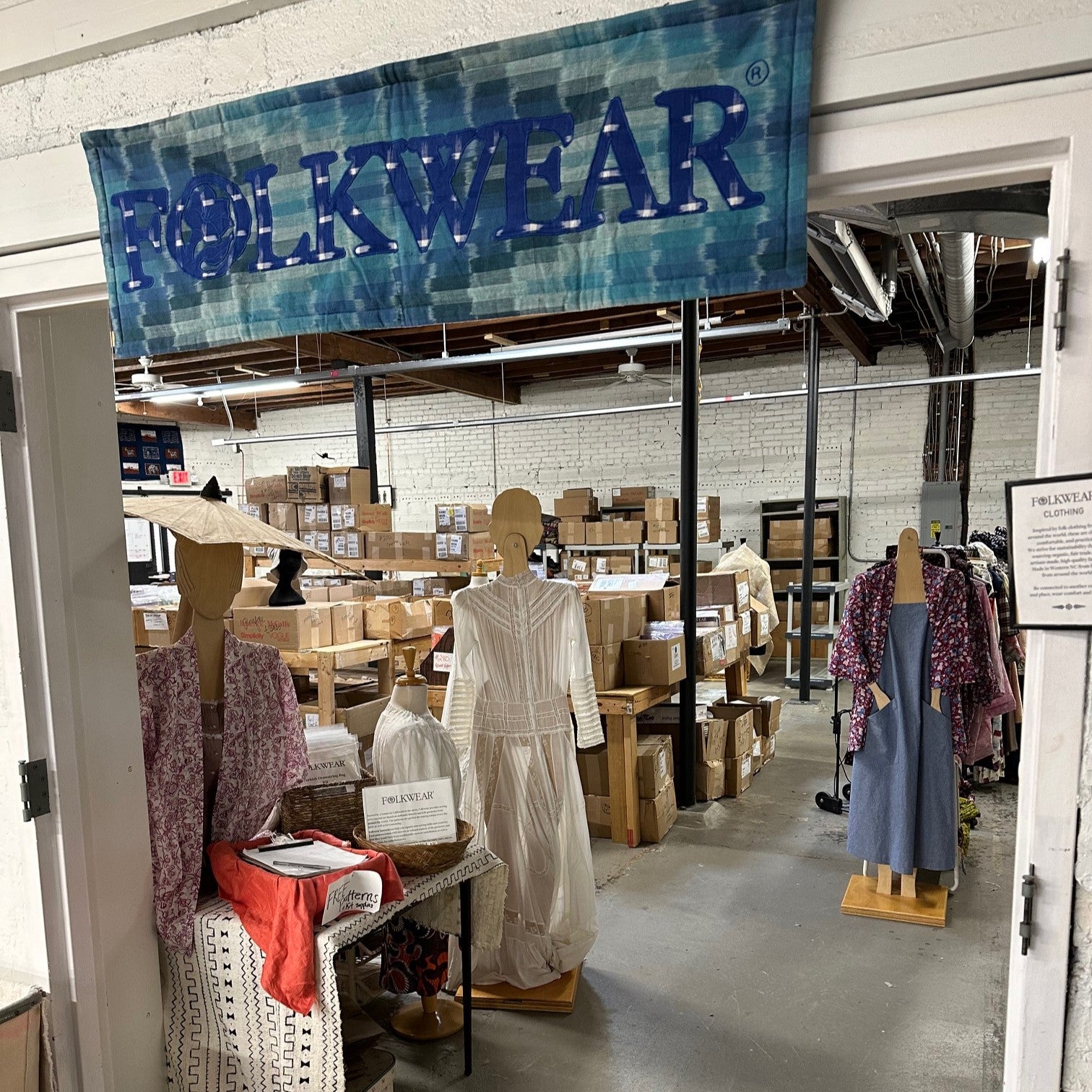 Entry to Folkwear with a blue fabric Folkwear banner above a room opening with manniquins wearing clothes and boxes in the background.