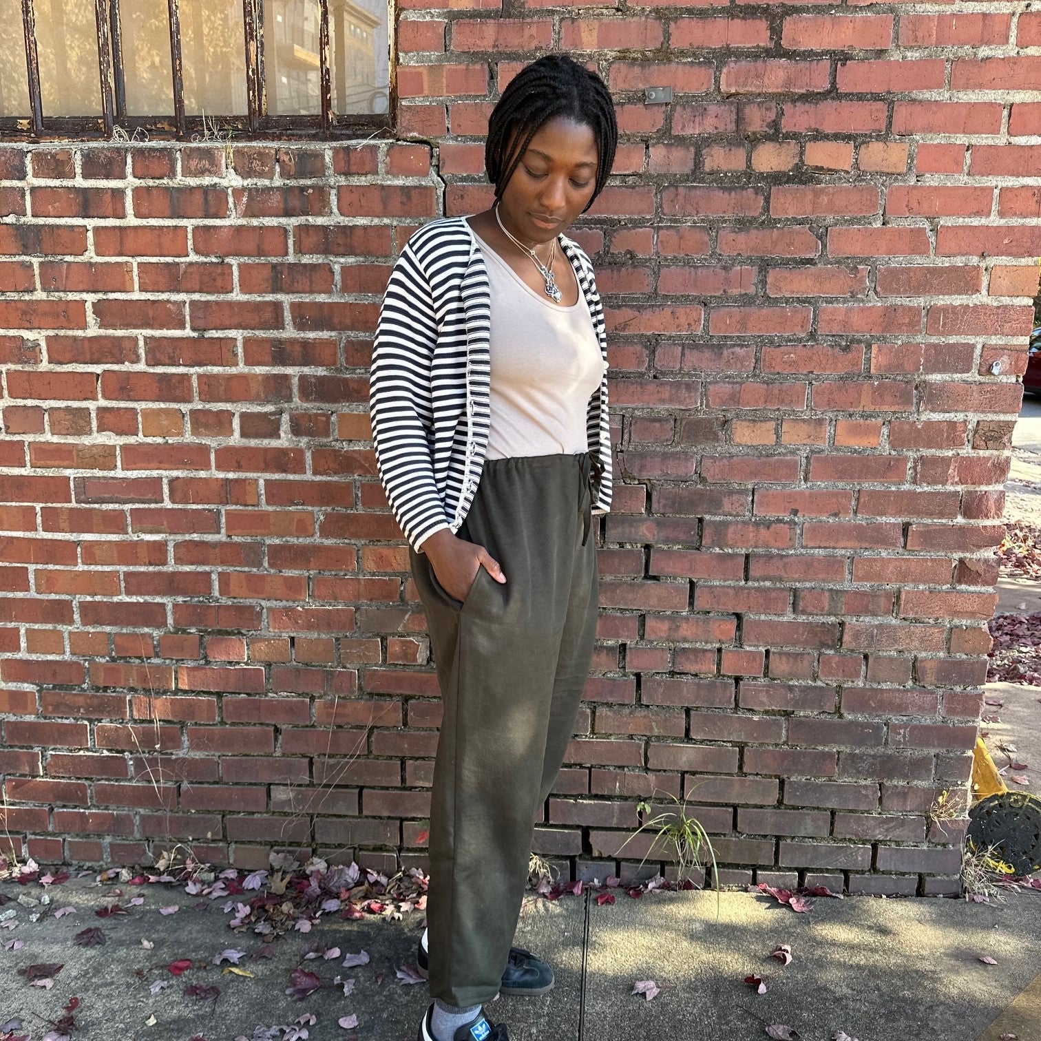 side view African women standing  in front of a brick wall wearing a black and white stripe cardigan and wearing 119 african saroulles in dark green with hands in her pockets.