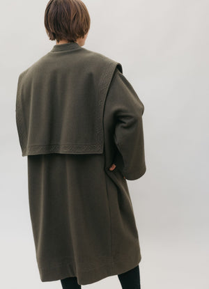 White woman standing with her hands in the pockets of a large dark olive green szur.  Her back is to the camera.