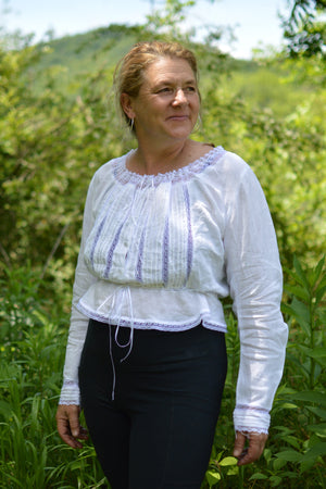 smiling middle aged white woman wearing a long sleeved camisole in white with white lace and black leggings with greenery in the background.