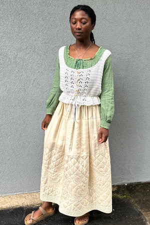 Black woman wearing a cream quilted skirt with a green blouse and white crocheted camisole on top - in front of a grey wall.