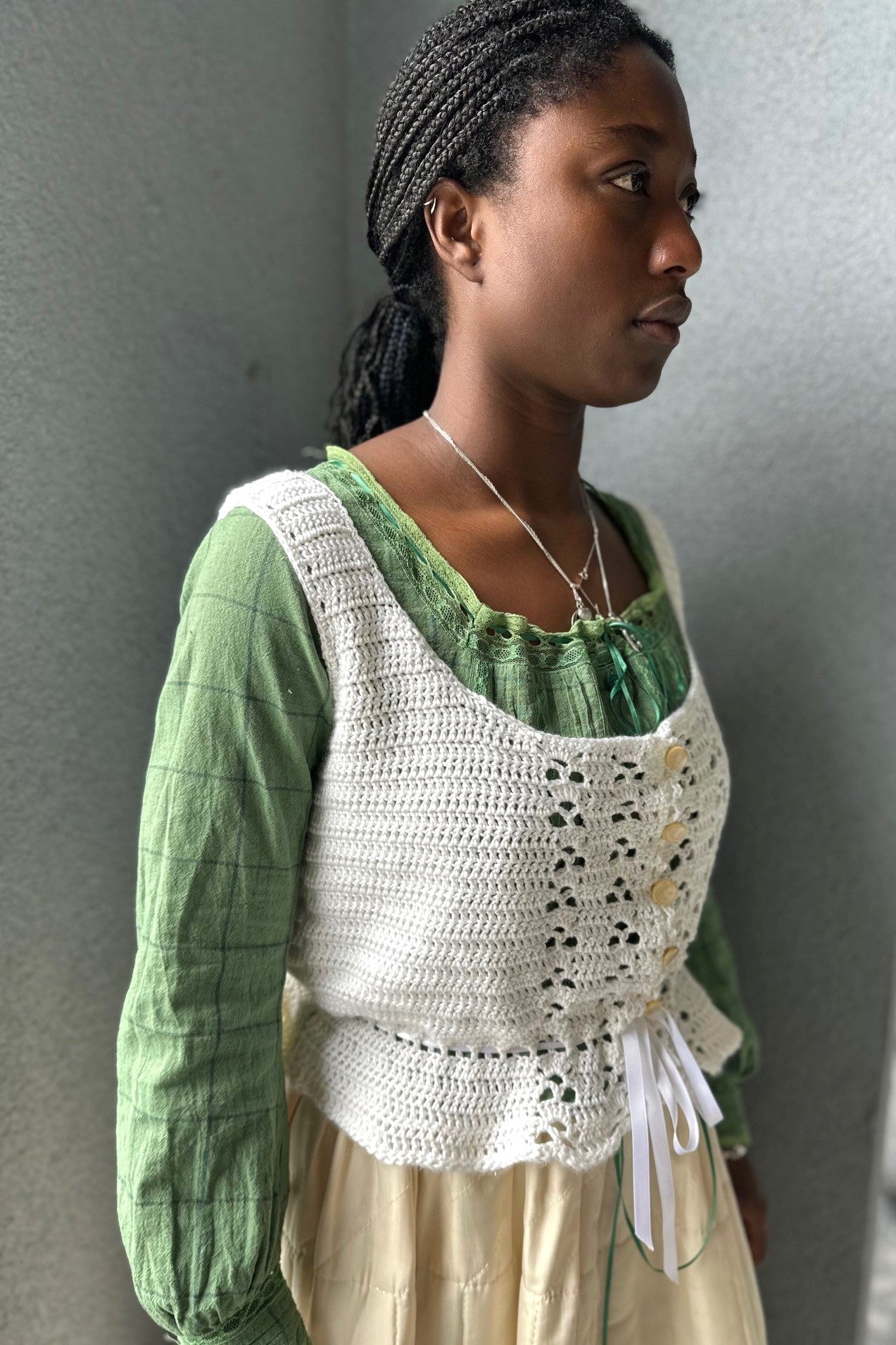 Black woman wearing a cream quilted skirt with a green blouse and white crocheted camisole on top - in front of a grey wall.