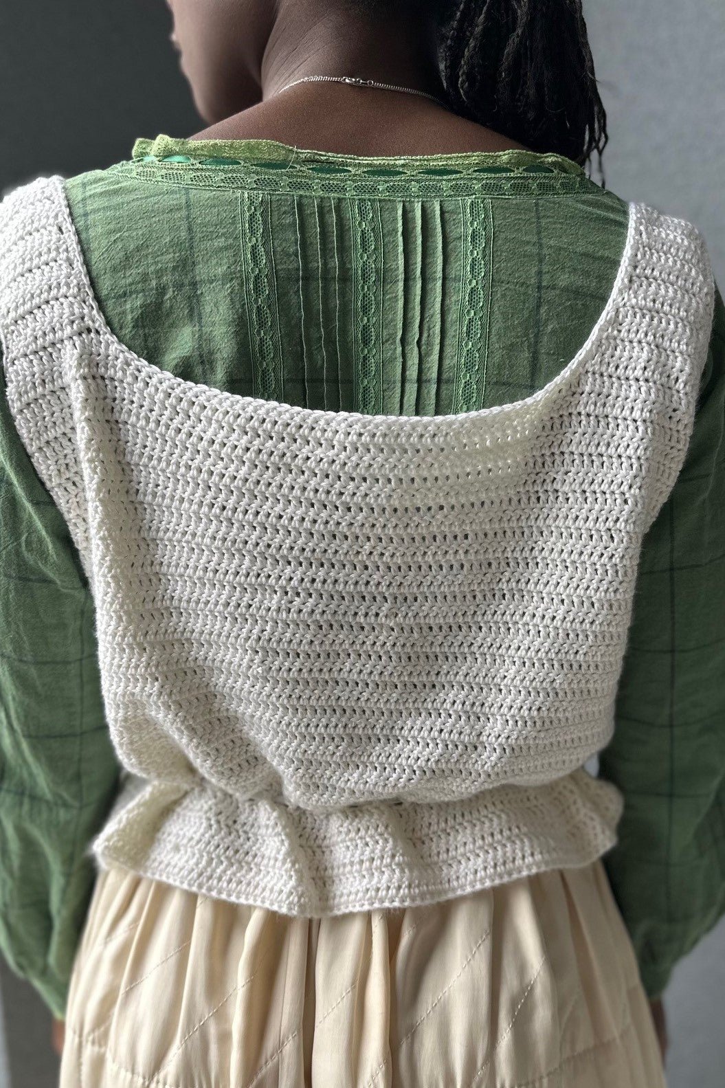 Black woman wearing a cream quilted skirt with a green blouse and white crocheted camisole on top - close up of the back - in front of a grey wall.