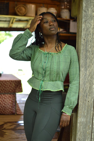 Front of African American woman leaning on a door frame with right hand moving her hair  wearing a green check 234 long sleeved version camisole and dark green athletic leggings.