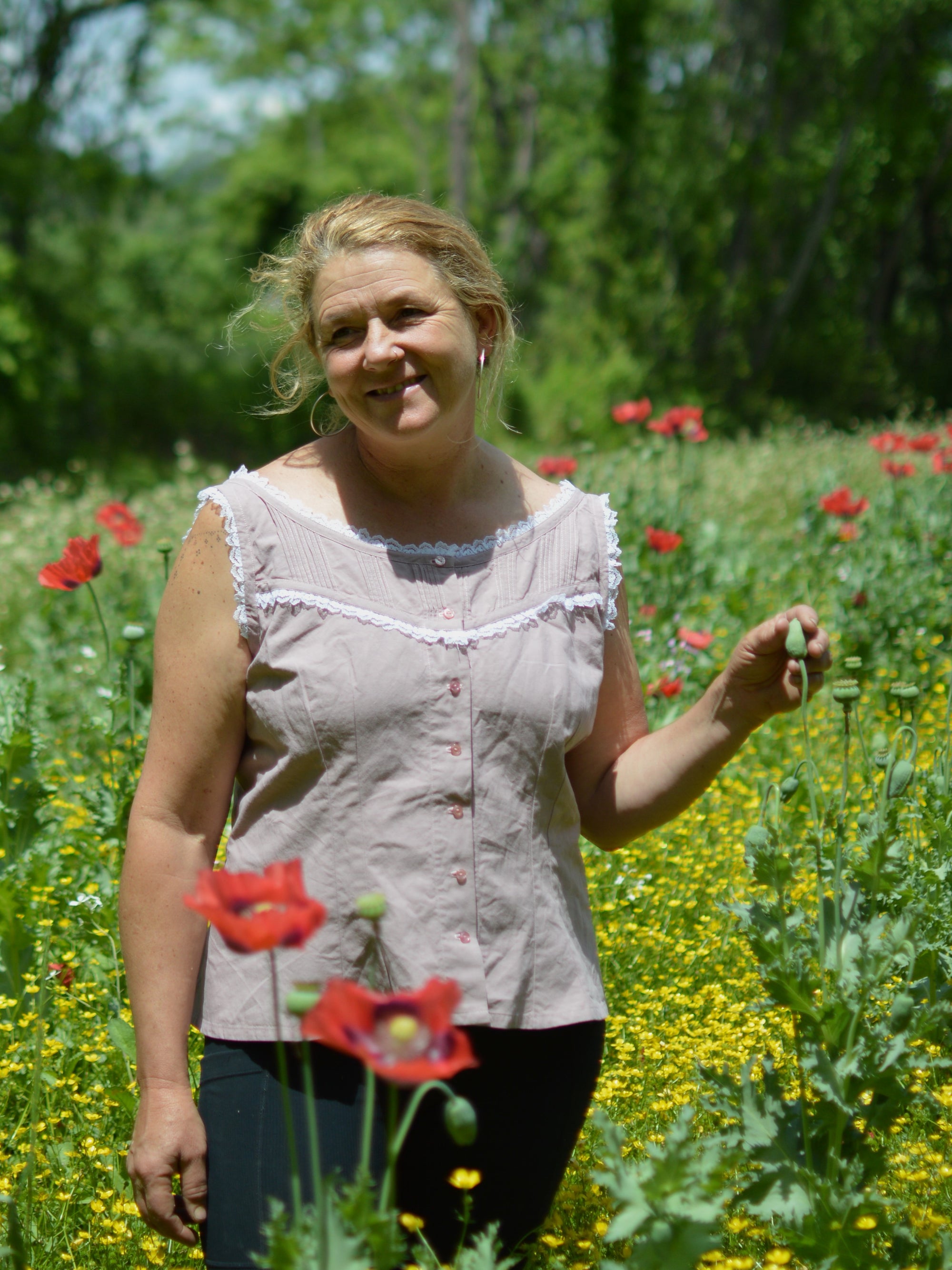 smiling middle aged white, blonde woman standing with left hand on a poppy bud wearing sleeveless camisole in pale pink with white ruffle lace and black athletic leggings she is standing in a field of red poppies.