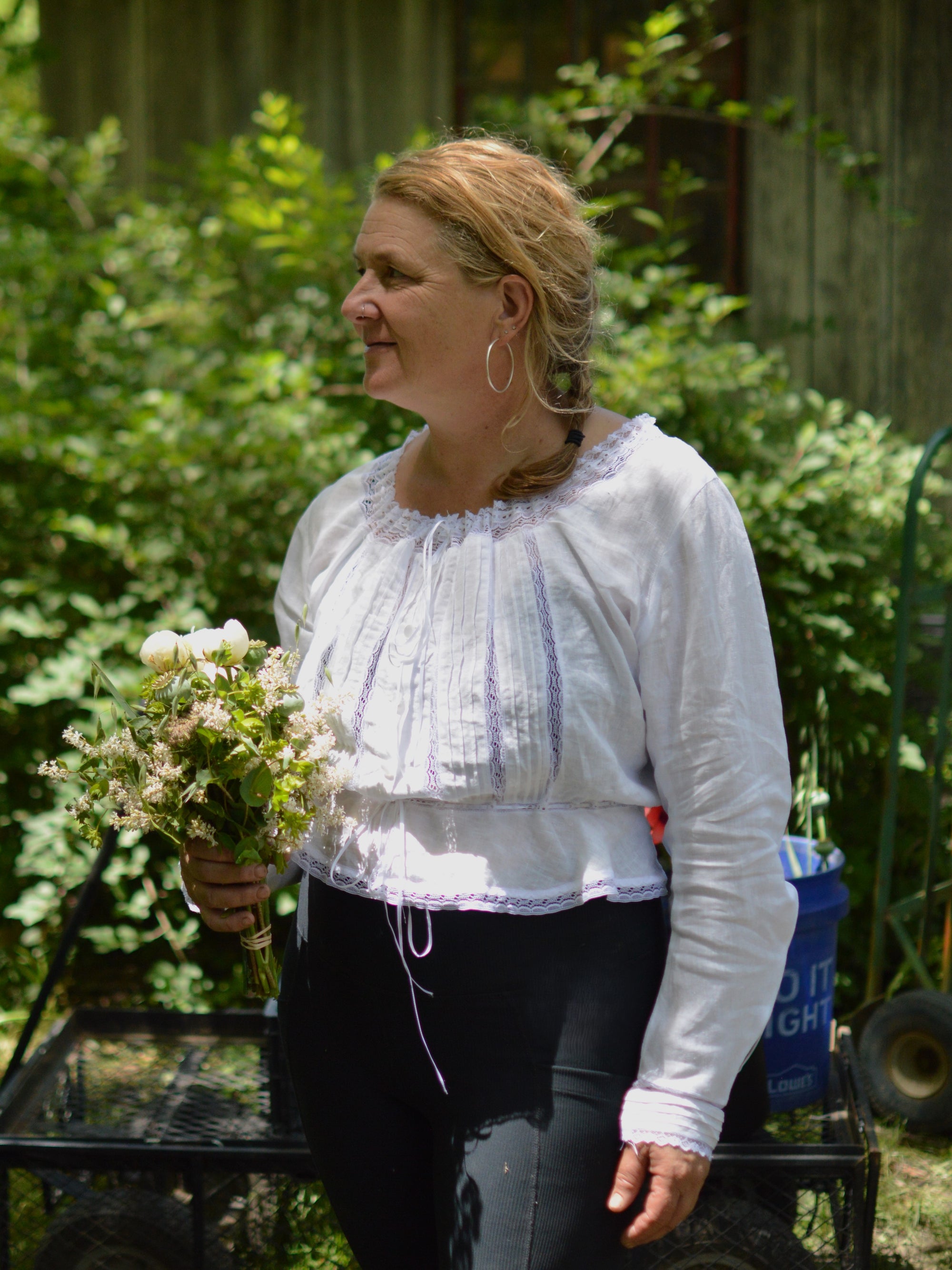 smiling middle aged white woman wearing a long sleeved camisole in white with white lace and black leggings with greenery in the background holding a bouquet.