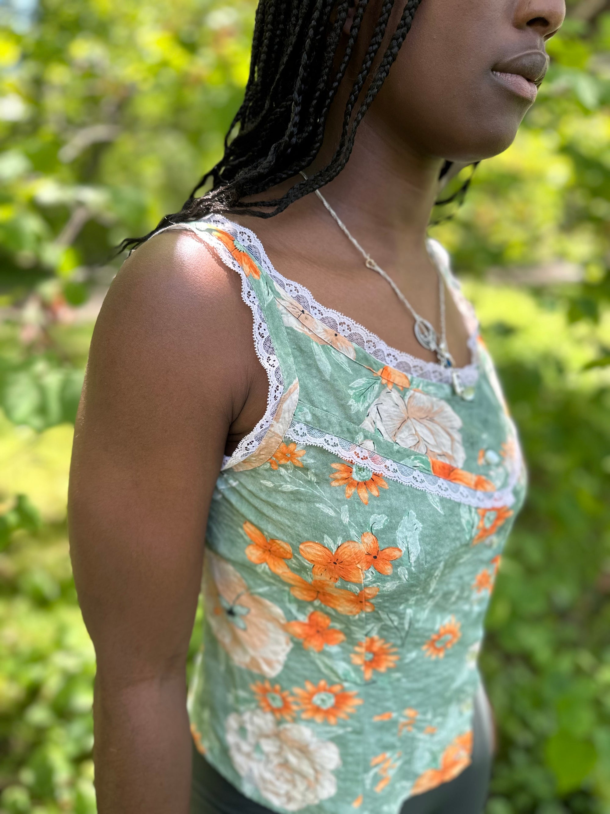 close up of side of sleeveless camisole in floral green and orange, worn by an african american woman with braided hair.