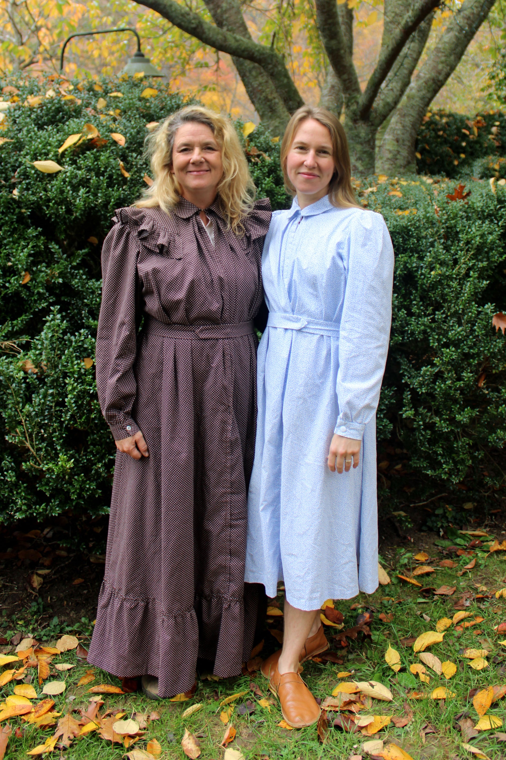 To women wearing a brown and blue Calico Day Dress, standing by a tree and bush outside.